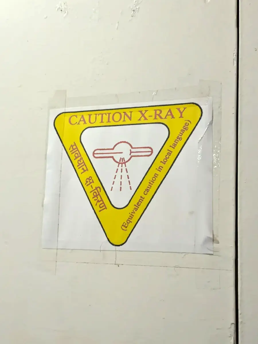 A yellow triangular sign on a white wall reads "CAUTION X-RAY" in English and Hindi, with a graphic of rays emitting from a device and text indicating equivalent caution in the local language.