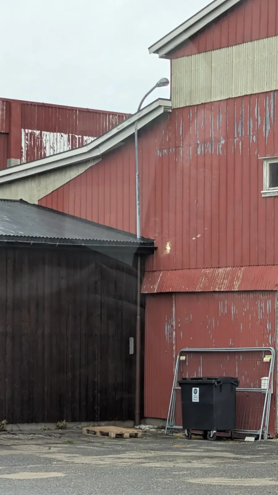 A weathered red and beige industrial building with peeling paint stands beside a dark wooden shed. A black trash bin and metal rack are visible in front, and a streetlamp is attached to the building.