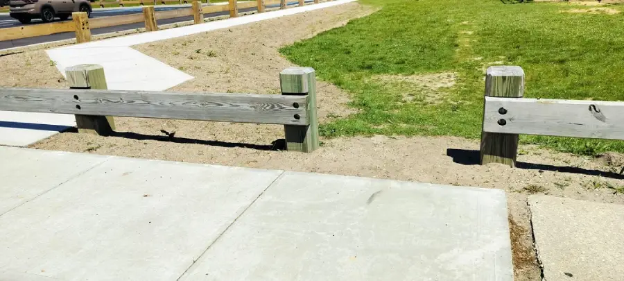 A sidewalk leads to a low wooden barrier with a gap, exposing dirt and grass beyond, making the path inaccessible for wheelchairs and strollers.