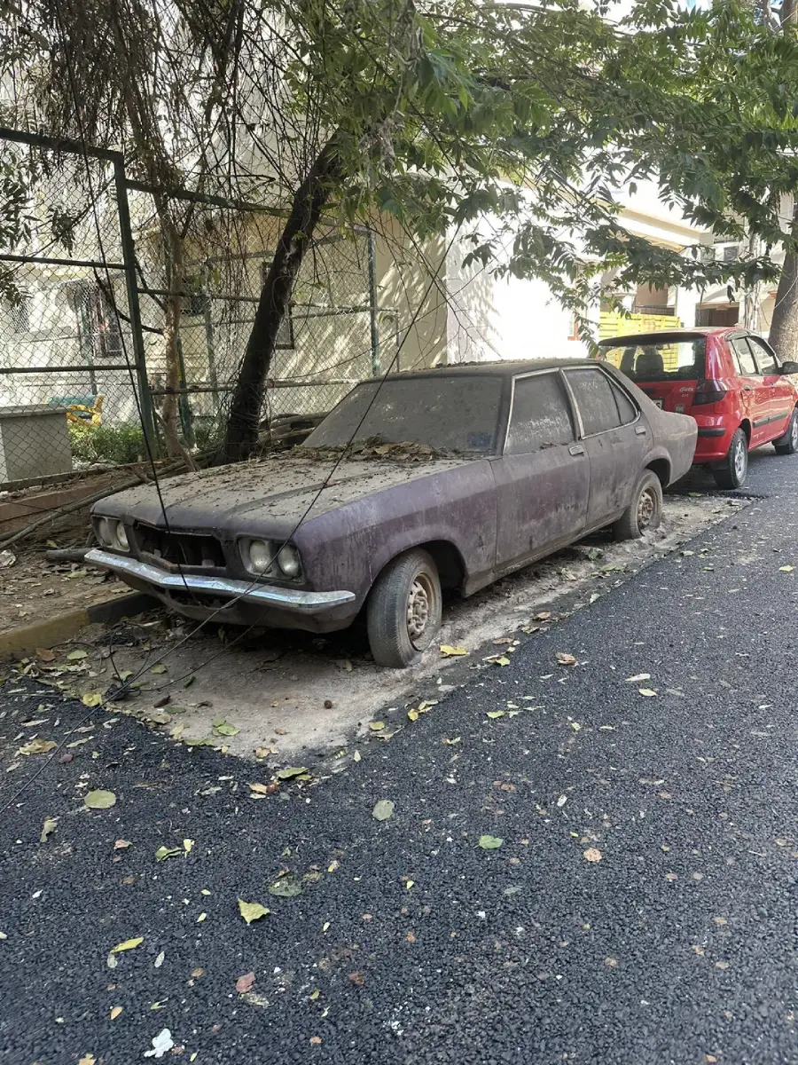 A rusted, dust-covered car with missing parts is abandoned on the side of a street, partly shaded by overhanging tree branches, with leaves scattered around and another car parked behind it.