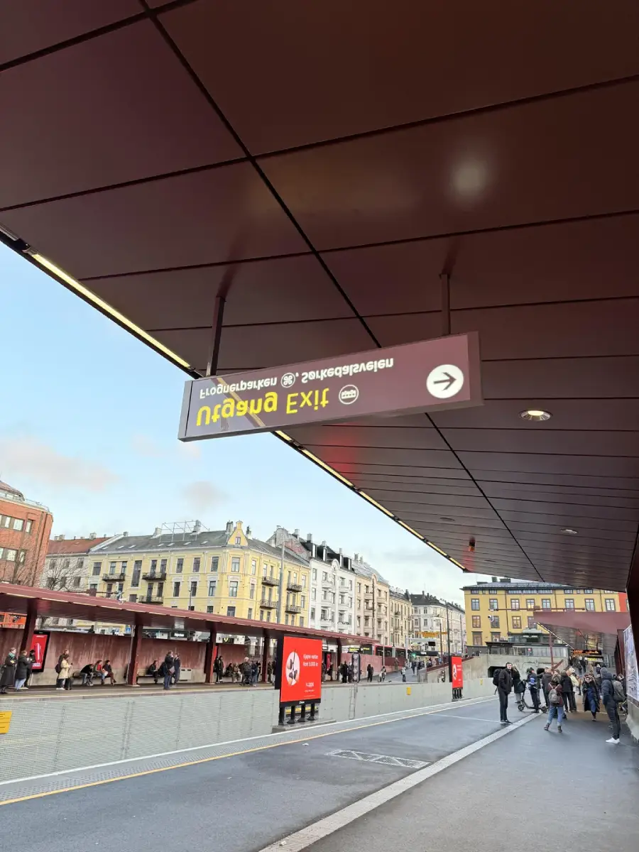 People walk along a city street next to a bus terminal. A large EXIT sign hangs from the ceiling, reflected upside down above. Classic European buildings and a partly cloudy sky are visible in the background.