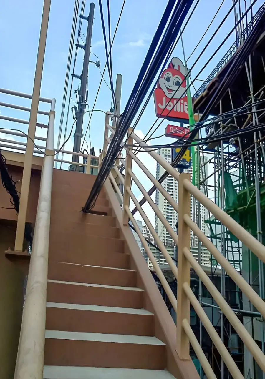 A beige outdoor staircase leads upward beside several power lines, with a Jollibee fast food sign visible in the background against a partly cloudy sky.