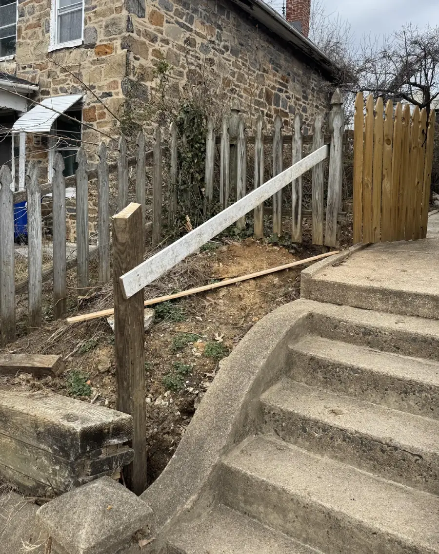 A concrete staircase leads up to a wooden fence, with a broken handrail leaning on a post beside uneven ground and a stone building in the background.