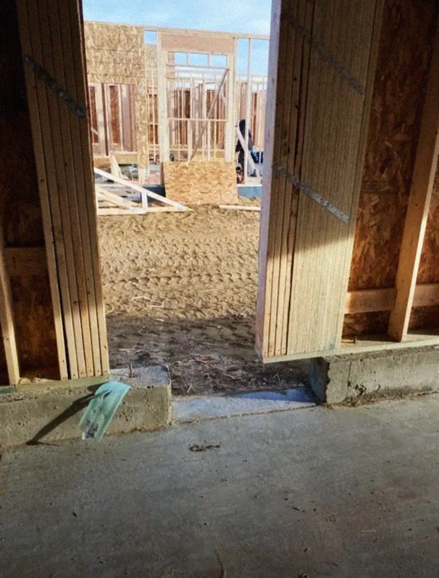 View from inside a building under construction, showing a doorway leading to a dirt area outside, surrounded by wooden framing and scattered construction materials.