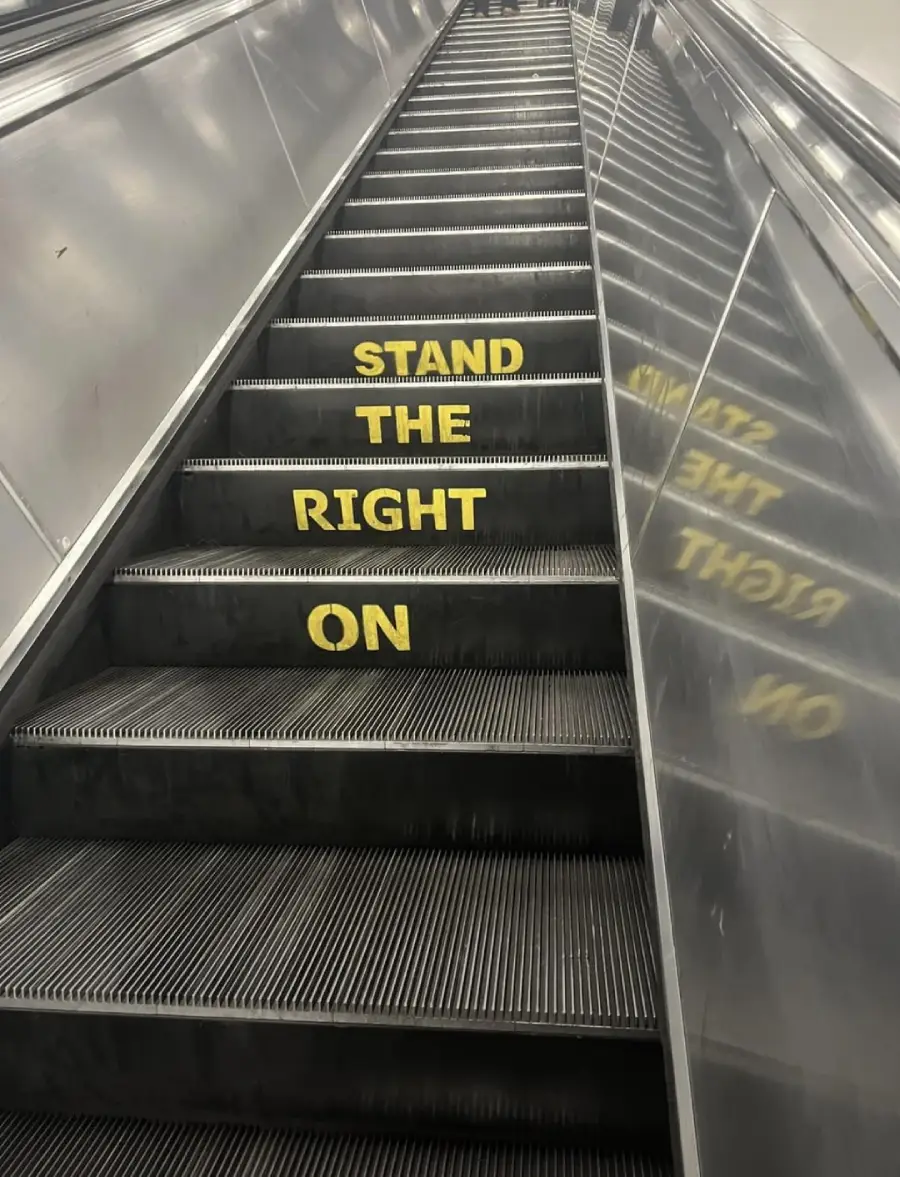 An escalator with yellow text on the steps reading “STAND THE RIGHT ON,” with a mirrored reflection showing the reversed text on the right side.