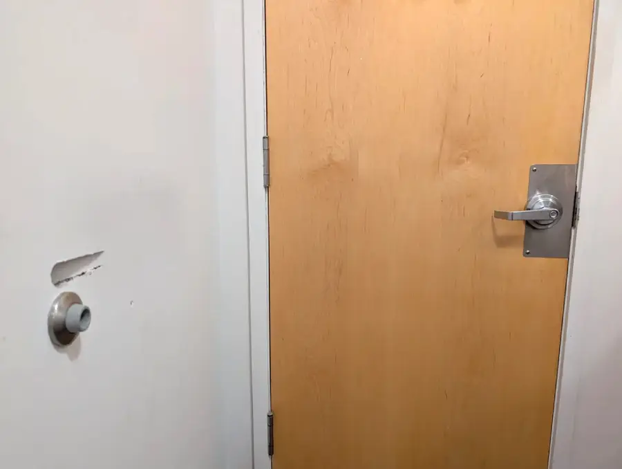 A wooden door with a metal handle is closed. The adjacent white wall has a doorknob-sized hole and scuff marks, suggesting the door handle has repeatedly hit and damaged the wall.