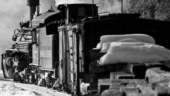 A black and white photo of a steam locomotive on snowy tracks with stacks of firewood and a layer of snow in the foreground. Steam rises from the train as it moves forward through a wintry landscape.