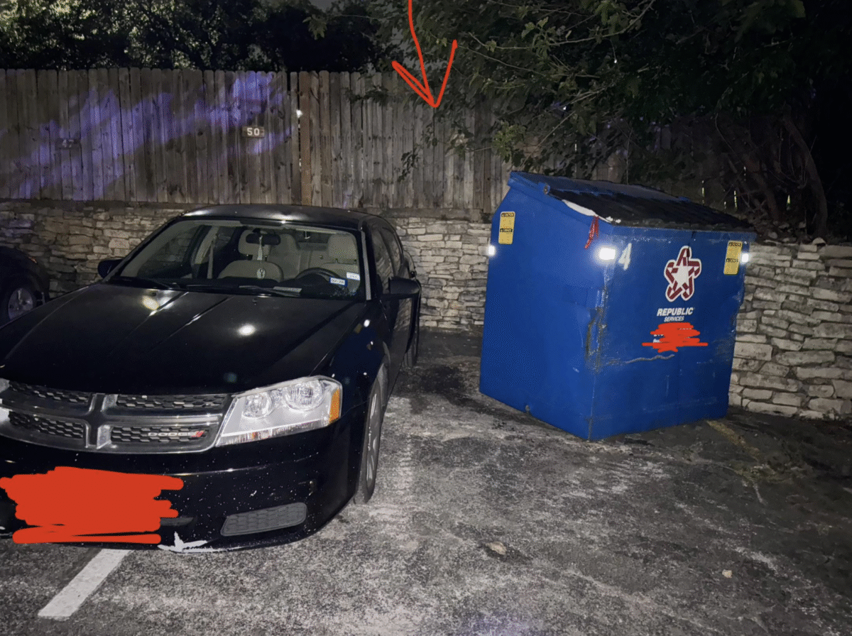 A black car is parked partly over the line and next to a blue dumpster in a dimly lit parking lot at night. A red arrow above points toward tree branches overhanging the area.