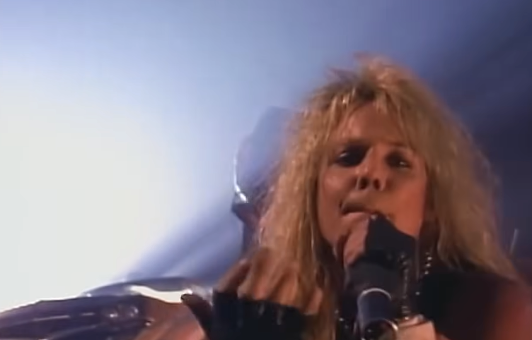 A long-haired singer performs energetically on stage, holding a microphone close, with dramatic lighting and smoke effects in the background.
