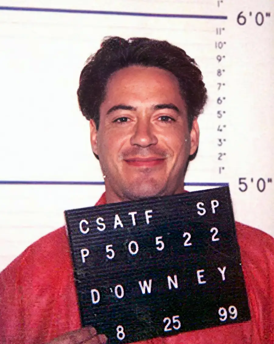 A man in an orange shirt smiles while holding a black sign with white letters and numbers in front of a height chart, as in a mugshot.