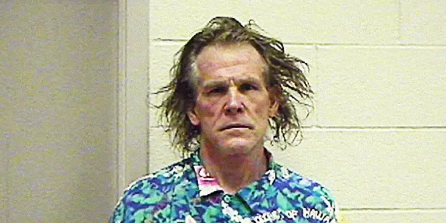 A middle-aged man with messy hair wearing a colorful floral shirt stands against a beige cinder block wall, looking directly at the camera with a serious expression.