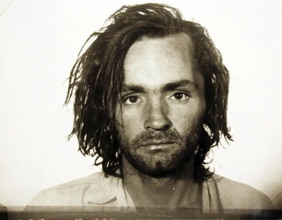 A black and white portrait of a man with long, messy hair and a scruffy beard, wearing a light-colored shirt, looking directly at the camera with a neutral expression.