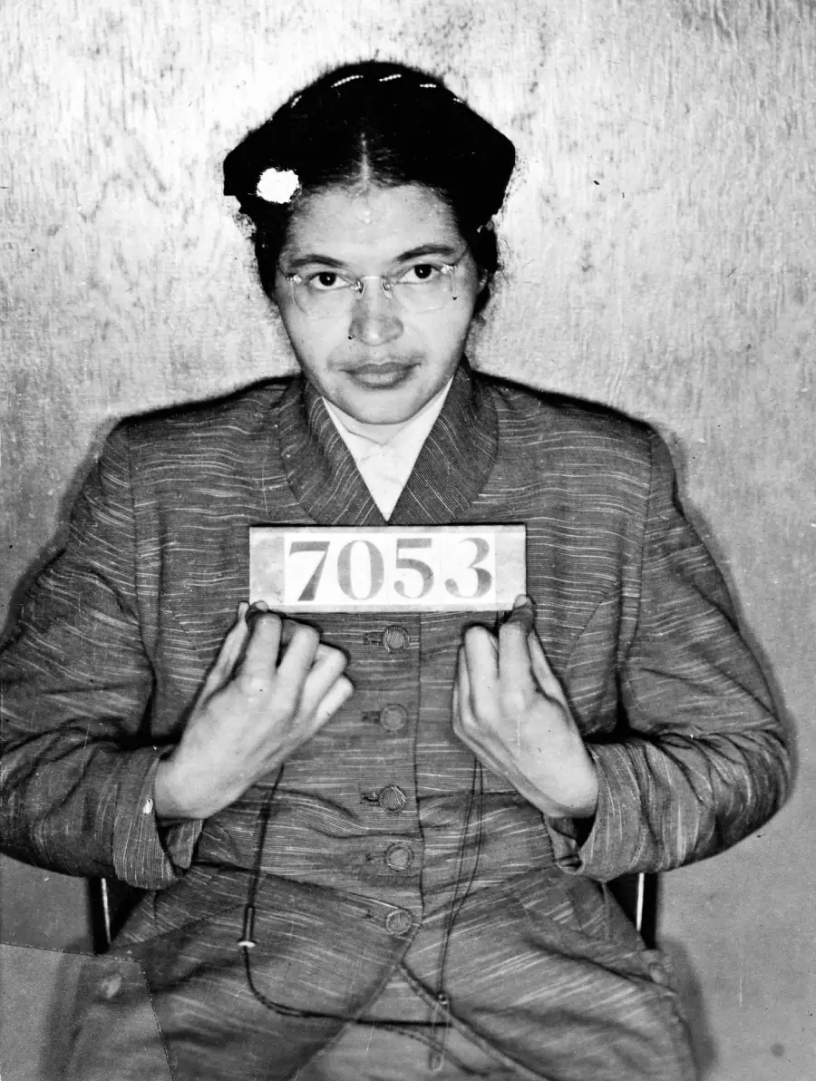 Black-and-white photo of a woman with glasses sitting against a plain wall, wearing a striped jacket and holding a numbered sign "7053" in front of her chest, as if for a mugshot.