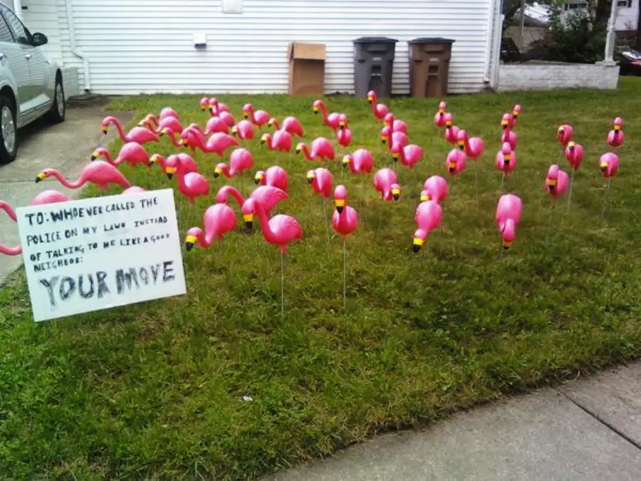 A lawn is covered with numerous pink plastic flamingos. A white sign reads: "To: Whoever called the police on my lawn instead of talking to me like a good neighbor: YOUR MOVE.
