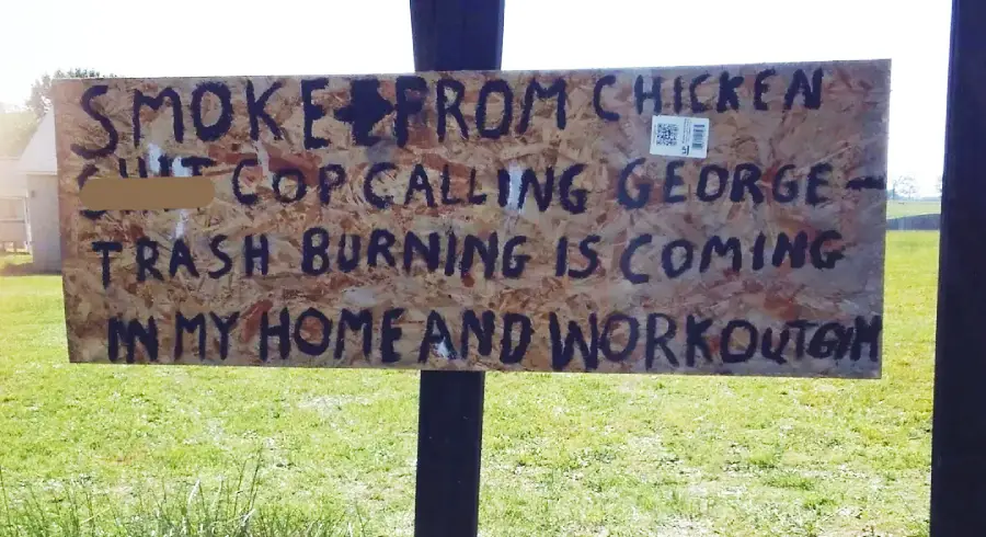 A plywood sign with black painted letters reads: “SMOKE FROM CHICKEN SH*T, COP CALLING GEORGE, TRASH BURNING IS COMING IN MY HOME AND WORKOUT.” The sign is outdoors on a grassy lawn.
