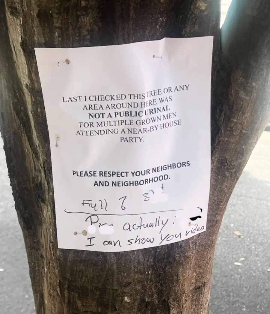 A paper sign on a tree reads: “Last I checked this tree or any area around here was not a public urinal for multiple grown men attending a nearby house party. Please respect your neighbors and neighborhood.” A handwritten reply underneath says, “Full of s**t. Actually I can show you video.”
