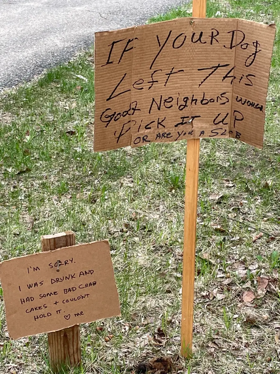 Two handwritten cardboard signs on stakes in grass. The larger sign asks dog owners to pick up after their dogs. The smaller sign is a humorous "apology" note from the dog's perspective, blaming bad crab cakes.