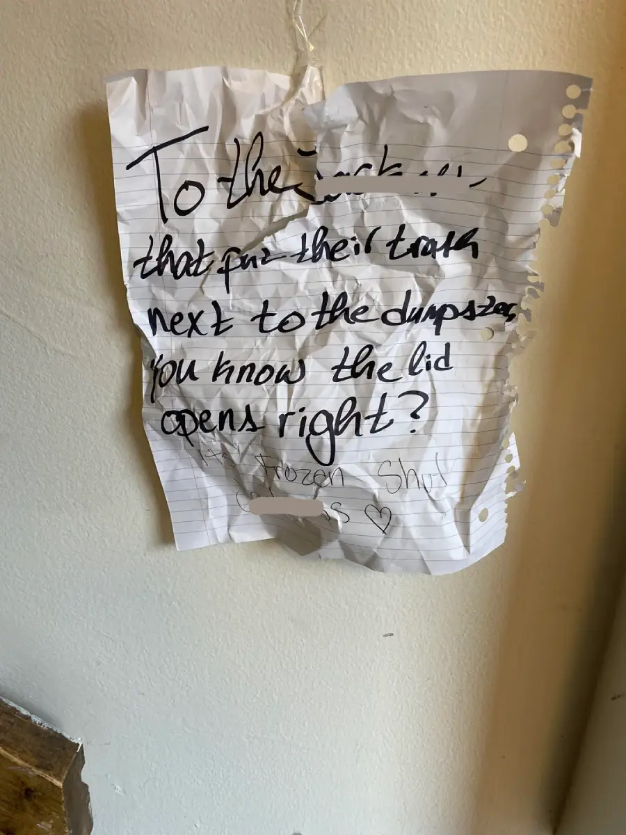 A crumpled, torn piece of lined paper taped to a wall displays a handwritten message reminding someone to put their trash in the bin and that the lid opens, with "Frozen Shrugs" signed at the bottom.