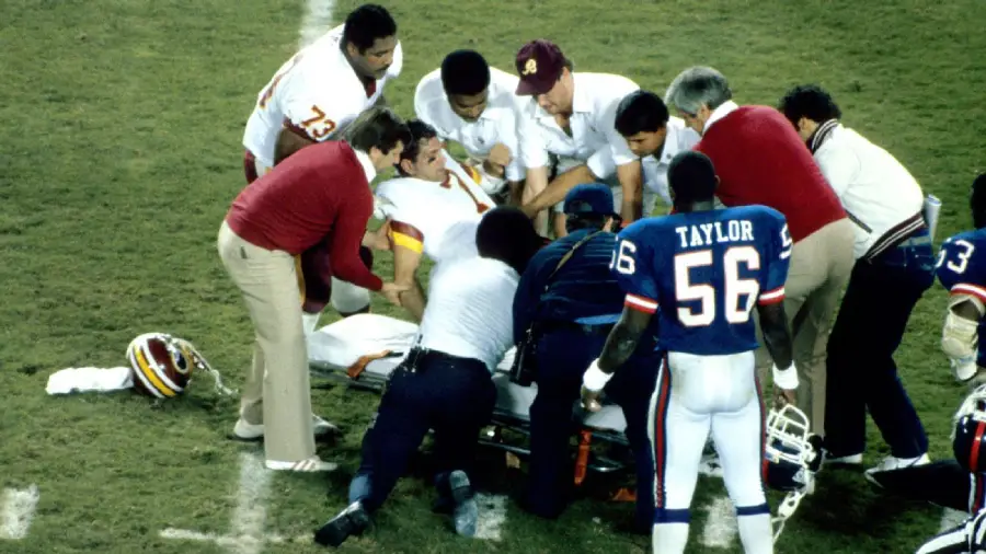 A football player in a white and red uniform lies on the field surrounded by trainers and staff, as medical personnel attend to him; a player in a blue uniform with number 56 stands nearby.