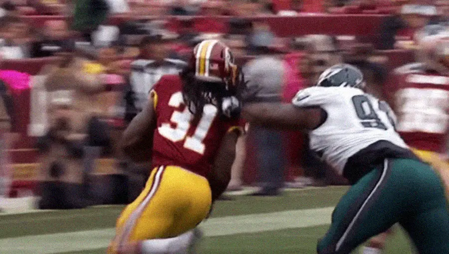 A football player in a burgundy and yellow uniform runs with the ball while a defender in a white and green uniform reaches out to tackle him, grabbing his face mask. The action takes place on a football field.