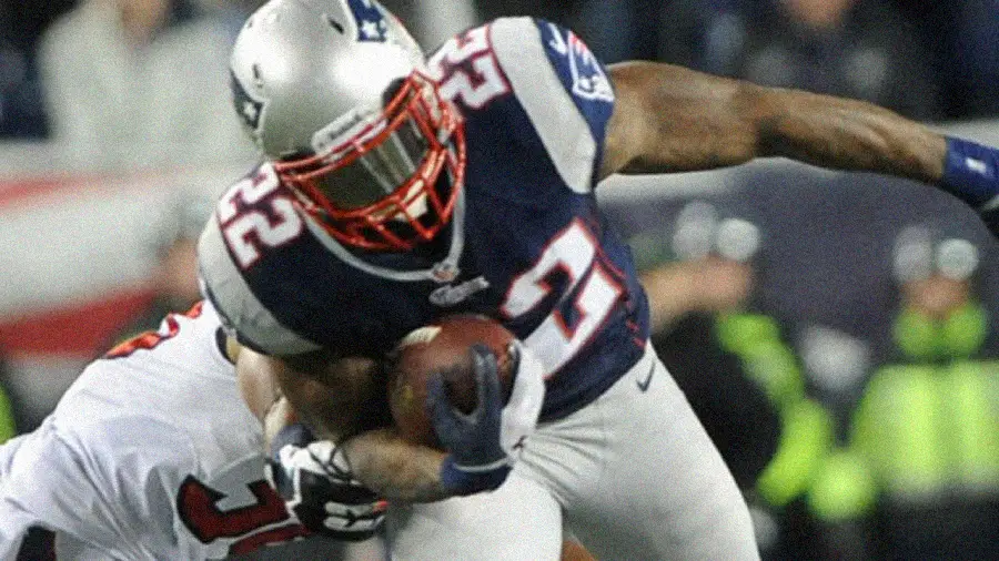 A football player in a New England Patriots uniform runs with the ball while being tackled by an opponent during a game.