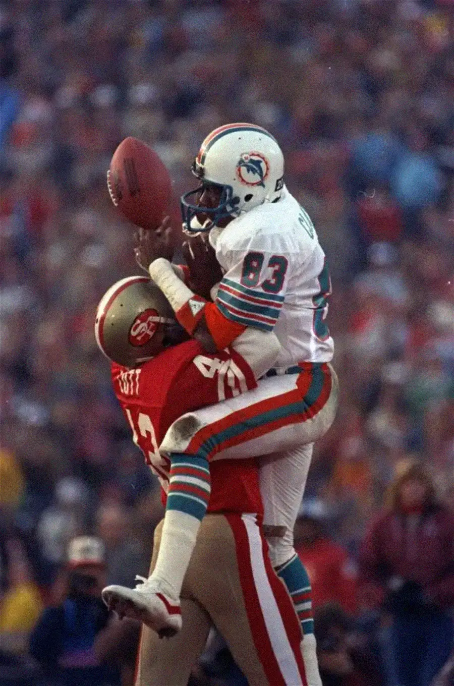 A Miami Dolphins player jumps to catch a football while closely defended by a San Francisco 49ers player during a game, with a blurred crowd in the background.