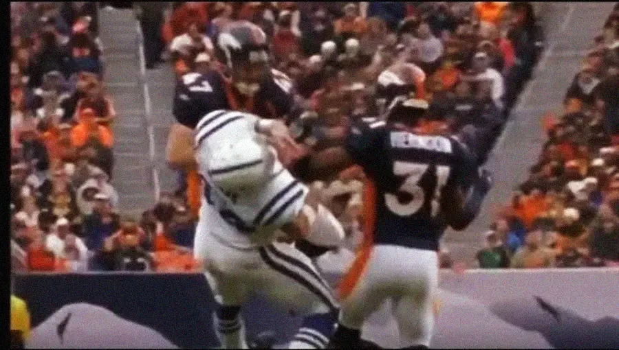 A football player in a white uniform is tackled hard by two players in dark uniforms during a game, with one player’s arm pushing the runner’s helmet back as the crowd watches in the background.