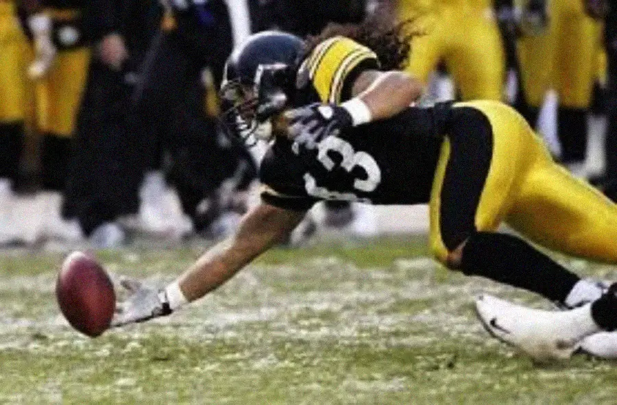 A football player in a black and yellow uniform dives forward with an outstretched arm, reaching for a loose football on the field during a game.