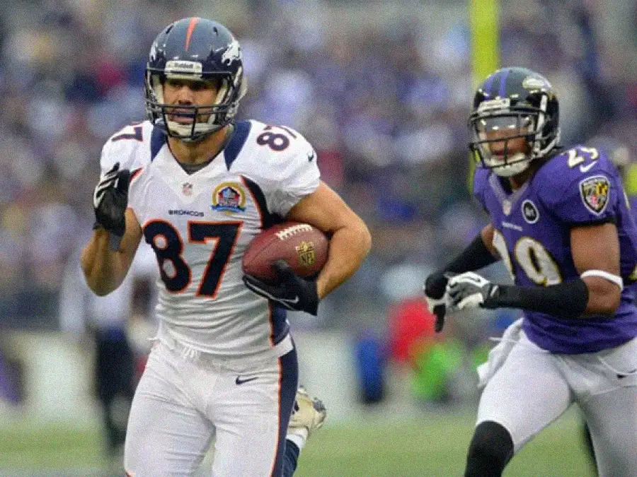 A Denver Broncos football player runs with the ball while being pursued by a Baltimore Ravens defender during an NFL game. Both players are in full uniform on a grassy field.
