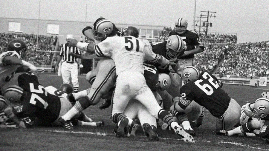 A black-and-white photo of a football game showing players from the Green Bay Packers and Chicago Bears in a crowded play at the line of scrimmage, with fans and referees visible in the background.