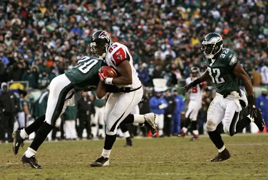 A football player from the Atlanta Falcons runs with the ball while being tackled by a Philadelphia Eagles defender, as another Eagles player approaches on a field with a crowd in the background.