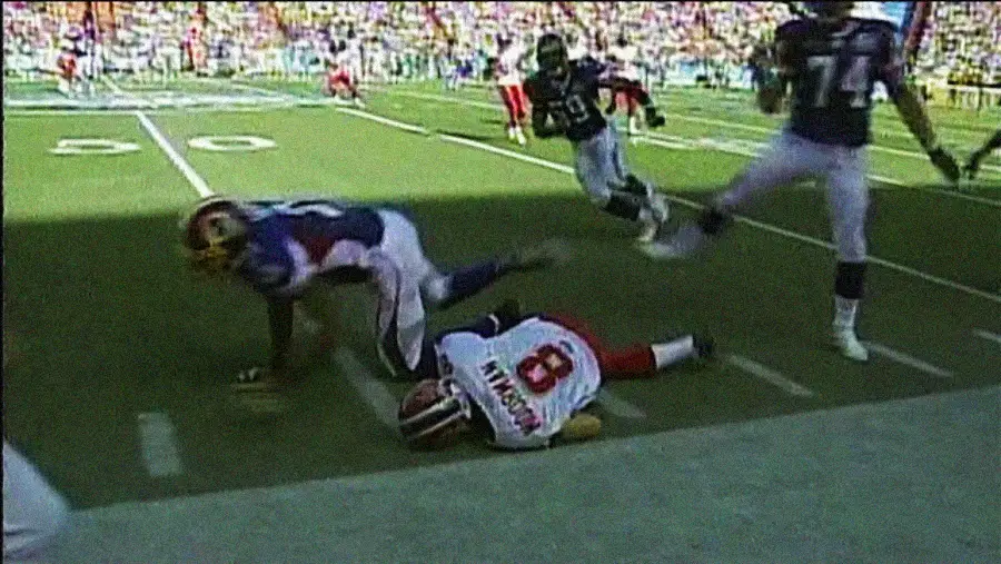 A football player in a white and red uniform lies face down on the field near the sideline as other players in blue and white uniforms move nearby during a game. The stands in the background are filled with spectators.