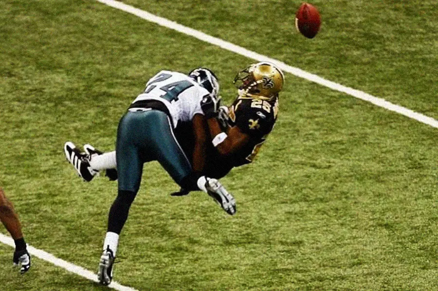 A football player in a green and white uniform tackles a player in a black and gold uniform, causing the football to fly out of his hands on a grassy field.
