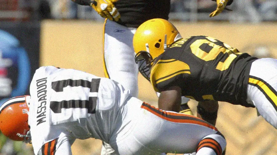 A football player in a white Cleveland Browns uniform, labeled "MASSAQUOI 11," bends forward as a player in a black and yellow Pittsburgh Steelers uniform attempts to tackle him during a game.