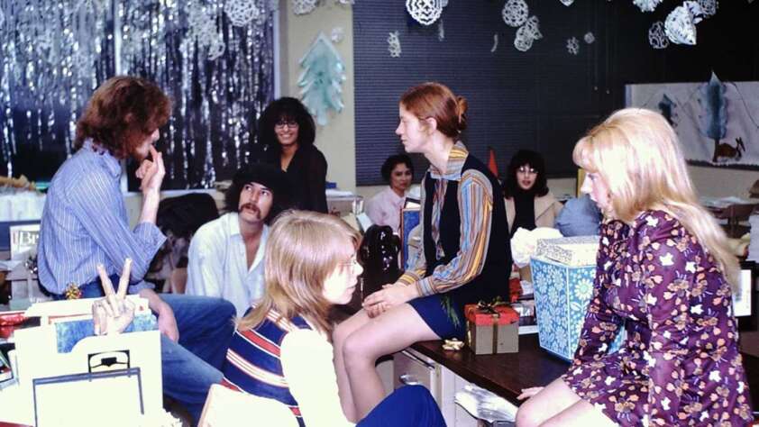 A group of young people sit and talk in a festive, decorated room with snowflakes and tinsel hanging from the ceiling. Wrapped gifts are on the table, and everyone appears relaxed and engaged in conversation.