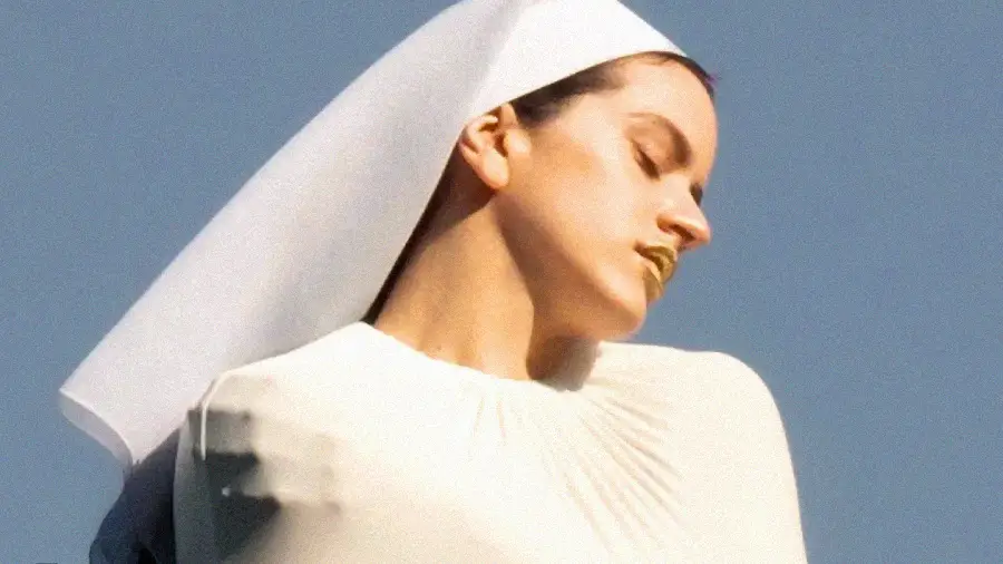 A person dressed in white with a white headpiece poses against a blue sky, eyes closed and head turned, with gold lipstick visible.