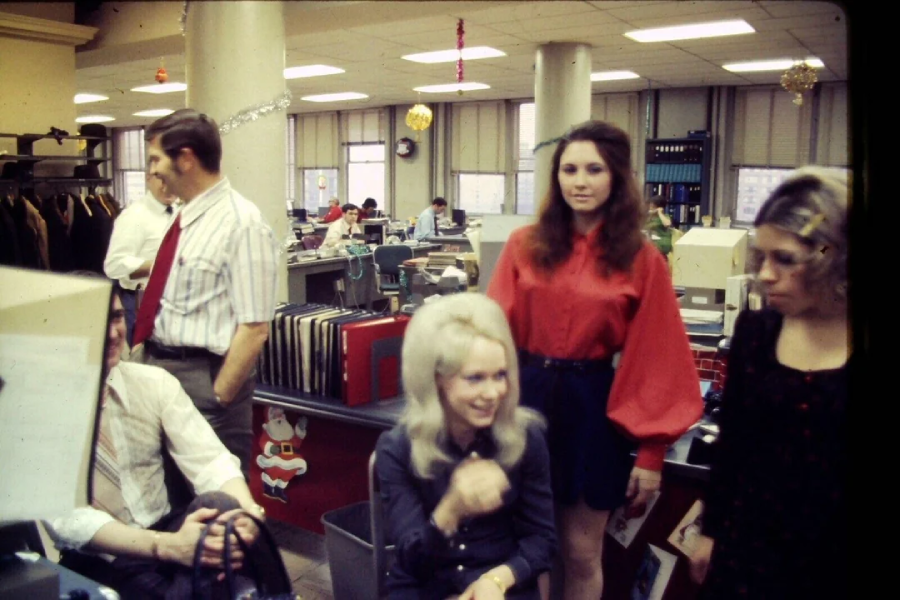 Several people are gathered in a busy office decorated with holiday ornaments. Three women are in the foreground, one with blonde hair seated and smiling, while a man in a shirt and tie stands nearby.