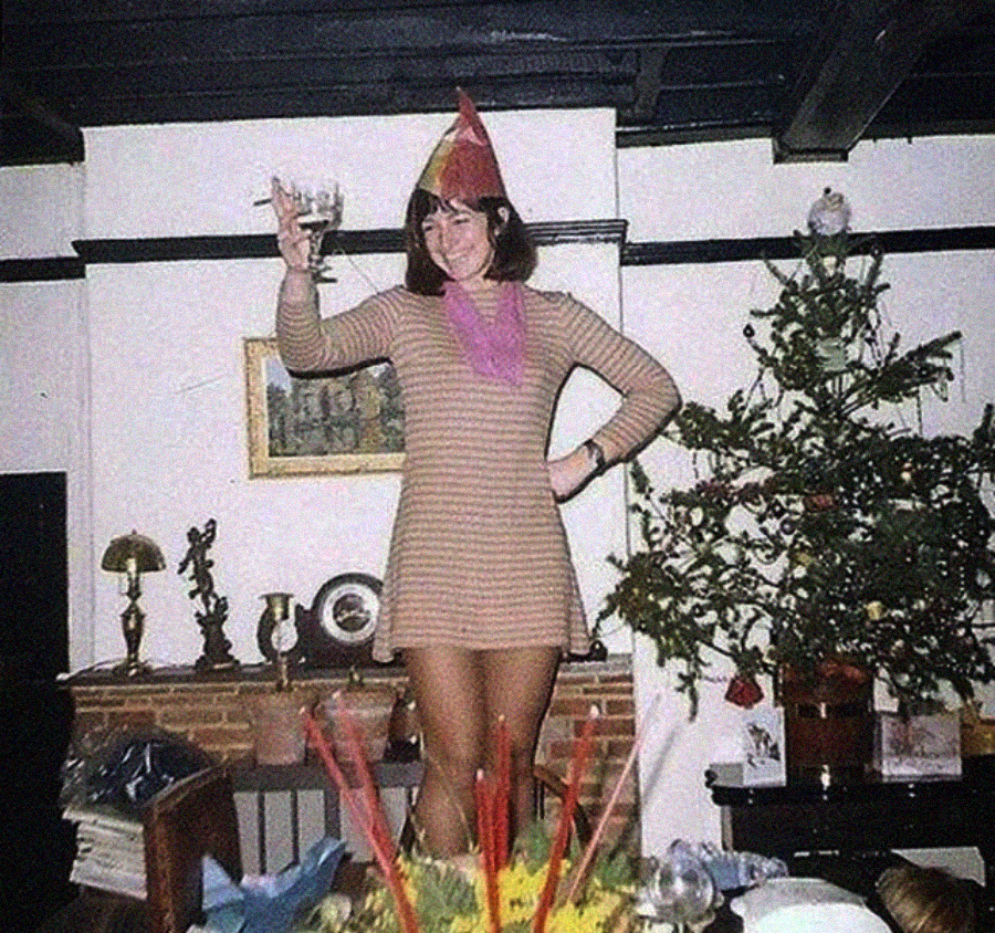 A woman in a striped dress and party hat stands on a table, raising a glass and smiling. There is a Christmas tree and festive decorations in the background.
