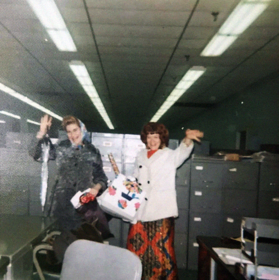 Two women smiling and waving inside an office with fluorescent lights, filing cabinets, and desks. One holds a large decorated bag and both are dressed in colorful, vintage-style clothing.