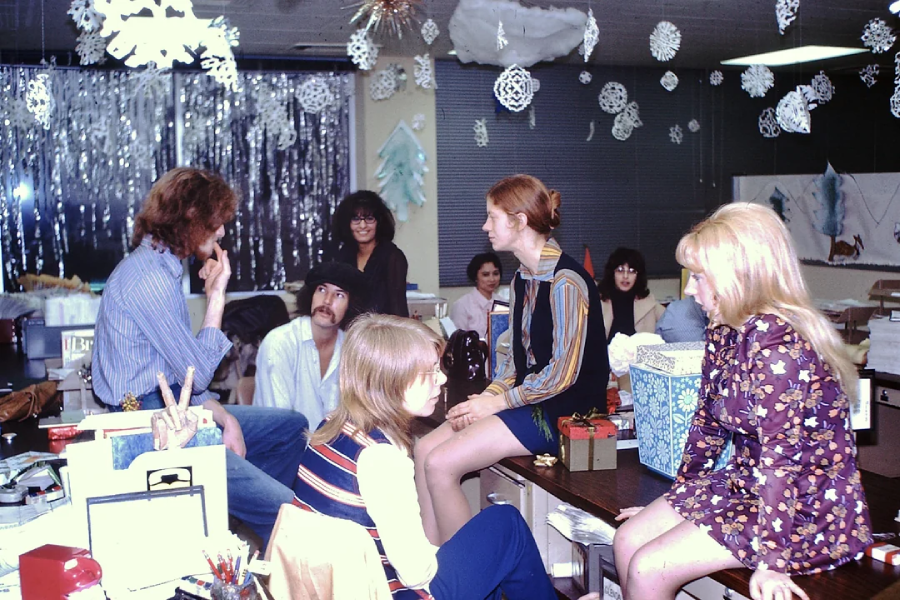 A group of young people sit and chat in a decorated office space with snowflakes and tinsel, suggesting a holiday or winter celebration. Gift boxes and festive decor are visible on desks and in the background.