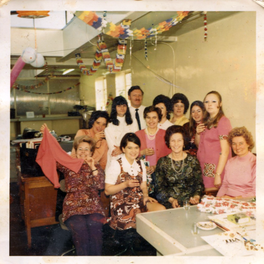 A group of people, mostly women, pose and smile at a lively indoor party with colorful streamers and paper chains hanging from the ceiling. Some are holding drinks, and a cake is on the table in front of them.