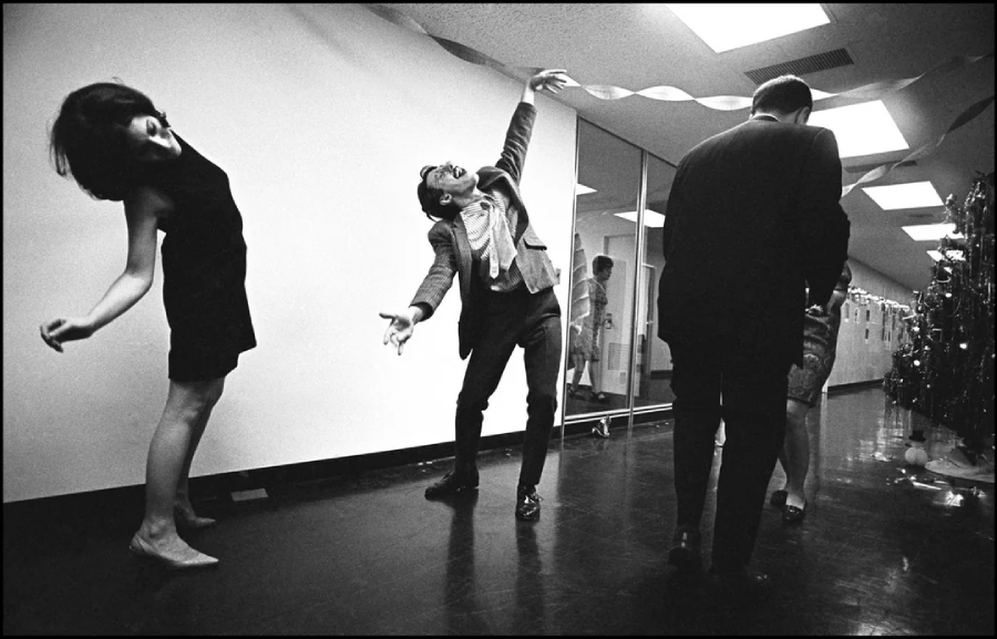 Three people dance and celebrate in a hallway decorated with streamers. One woman leans back with a smile, while a man with glasses poses playfully, arms outstretched. Two other people stand with their backs to the camera.