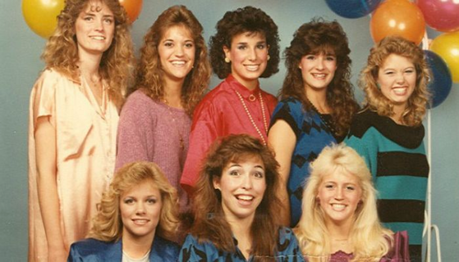 Eight women with big, curly 1980s hairstyles and colorful outfits pose together in front of a light blue backdrop with orange, purple, and yellow balloons in the background. They are smiling at the camera.