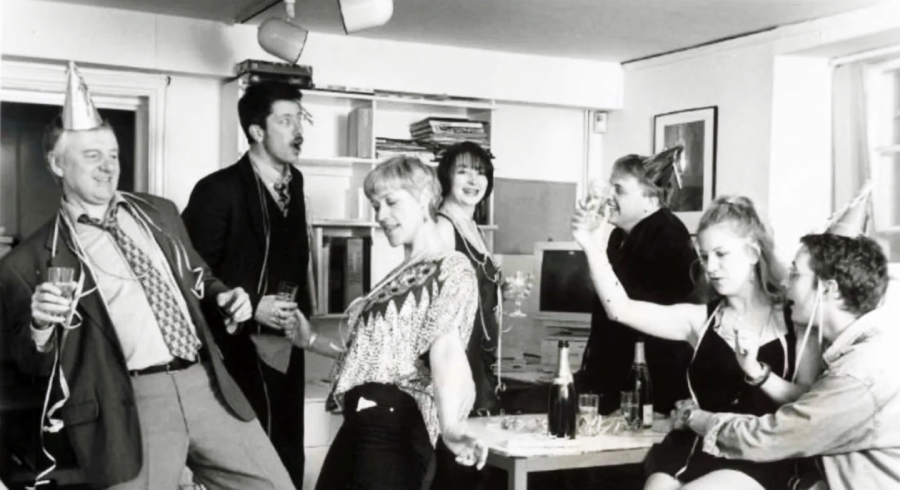 A group of people in party hats dance, laugh, and drink at an office party. Some hold glasses, while others pose and celebrate together in a lively, festive atmosphere.