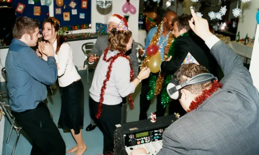 People in festive clothes and party hats dance and laugh at an indoor holiday party. A DJ plays music in the foreground, and decorations like tinsel and paper snowflakes hang around the room.