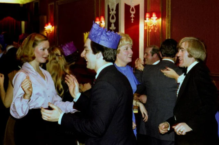A group of people wearing colorful paper crowns and formal attire are smiling, talking, and dancing together at a festive indoor party with red walls and warm lighting.