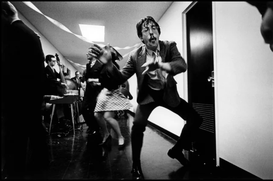 A man with messy hair energetically dances in a hallway decorated with streamers, while others dance and a small band plays instruments in the background. The atmosphere is festive and lively.