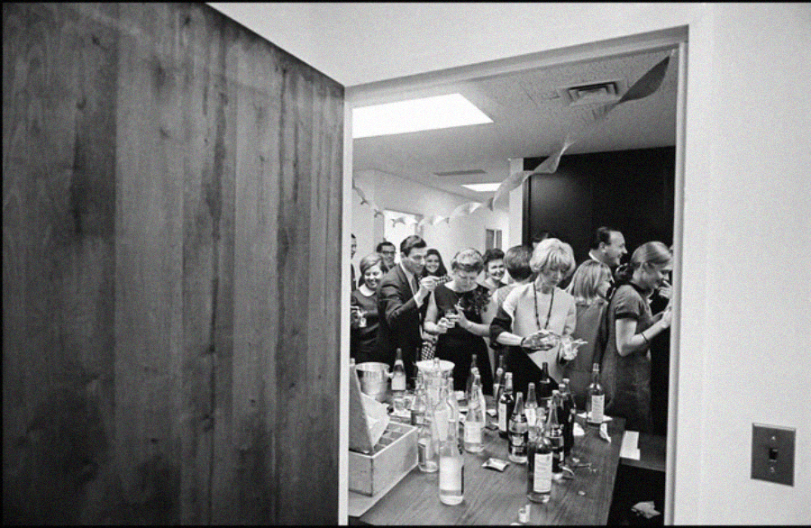 A group of people gather in an office room for a party, holding drinks and plates of food. Bottles and cups are on a table in the foreground. The scene appears lively and social.