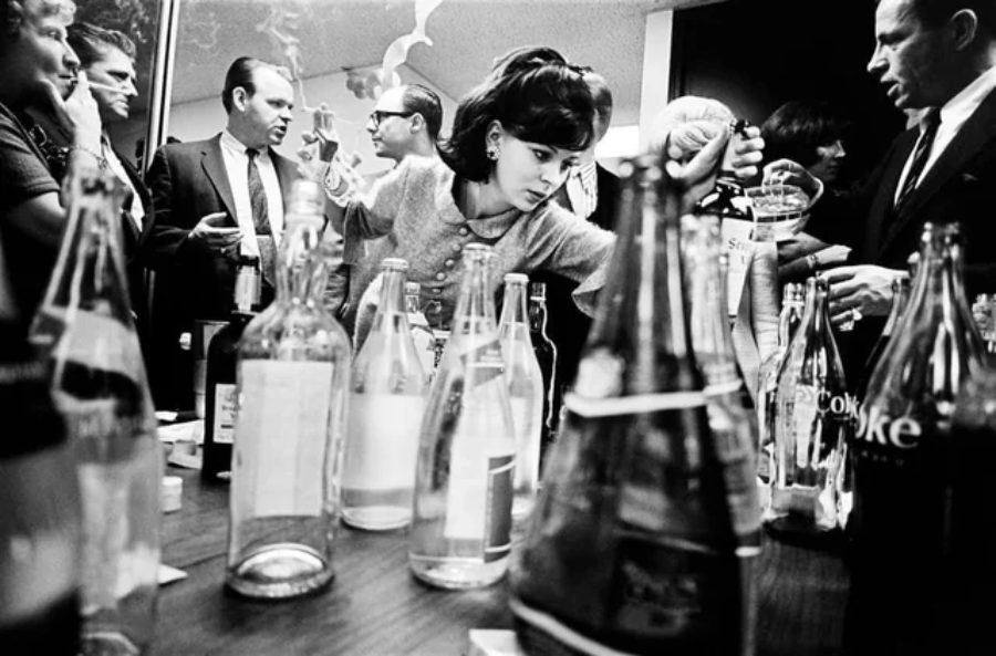 A woman reaches for a bottle among many glass bottles on a table as several people in business attire gather and interact in a busy room. The image is in black and white.
