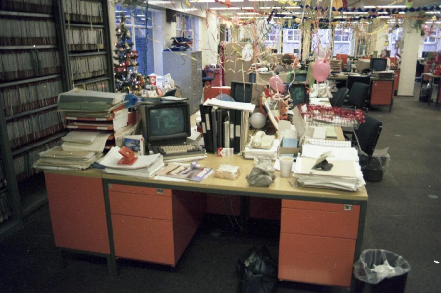 A cluttered office desk with papers, folders, and an old computer, surrounded by cubicles decorated with streamers and balloons. A decorated Christmas tree is visible in the background, suggesting a festive season.